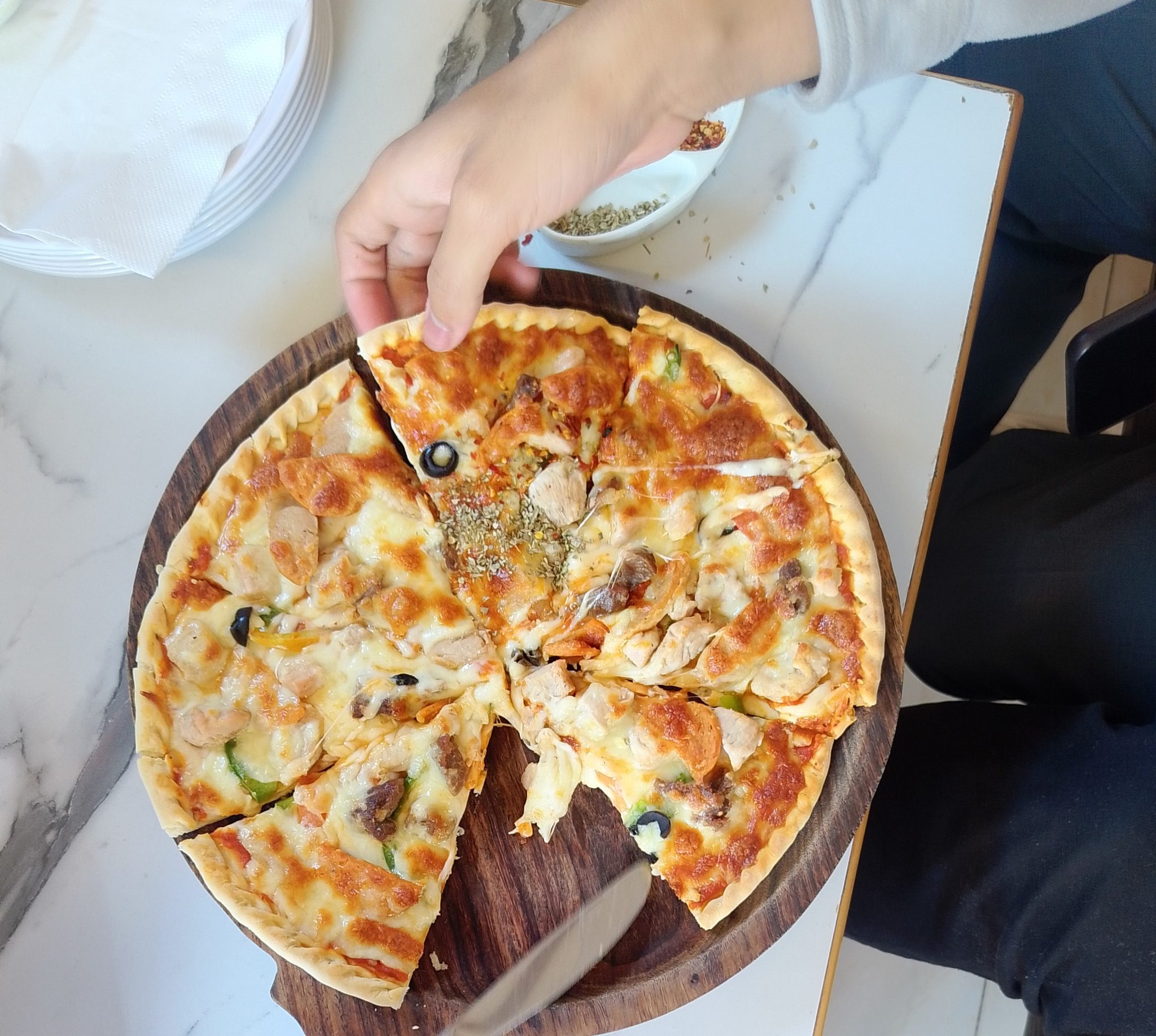 Ahşap tahtada karışık malzemeli pizza, bir el dilim alırken yakalanmış lezzetli an