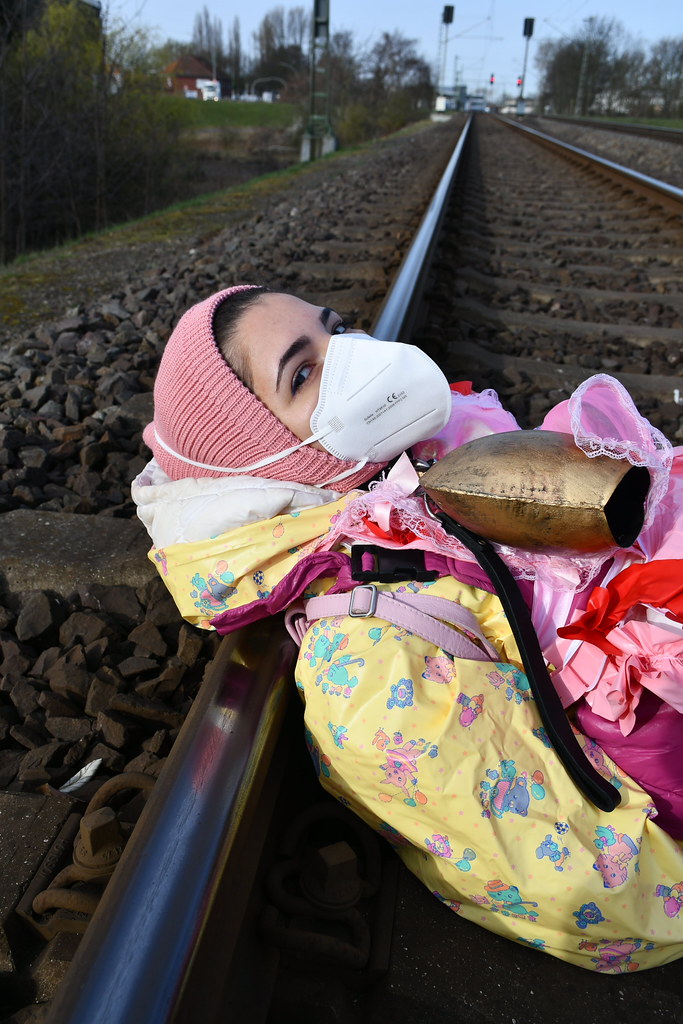 Maske ve Pembe Bereyle Tren Raylarına Yatmış Renkli Giysili Kadın Görüntüsü